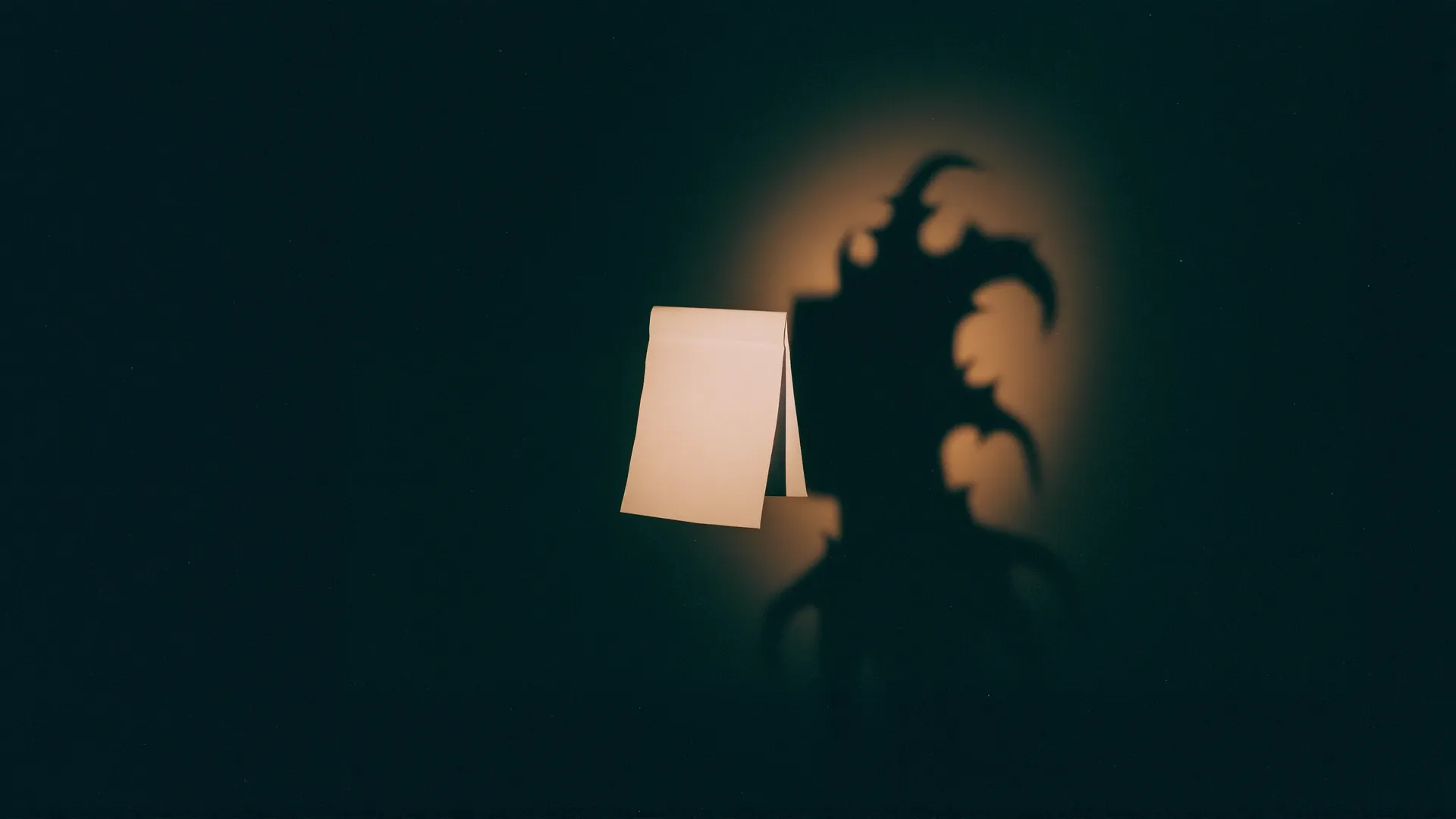 A plain pale sheet of paper standing in darkness, lit by a warm glow, casting a large clawed and winged monster-shaped shadow on the wall behind it that does not match the calm object making it