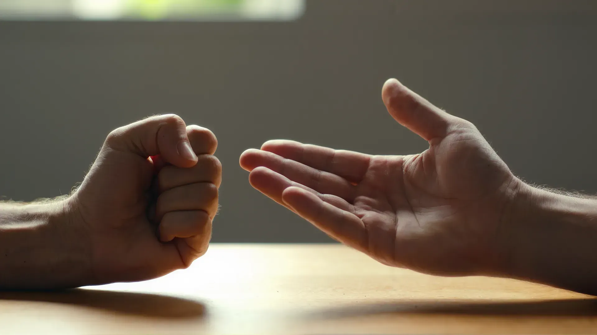 Close-up of one person's two hands on a wooden table — the left hand clenched into a tight fist, the right hand fully open, palm up