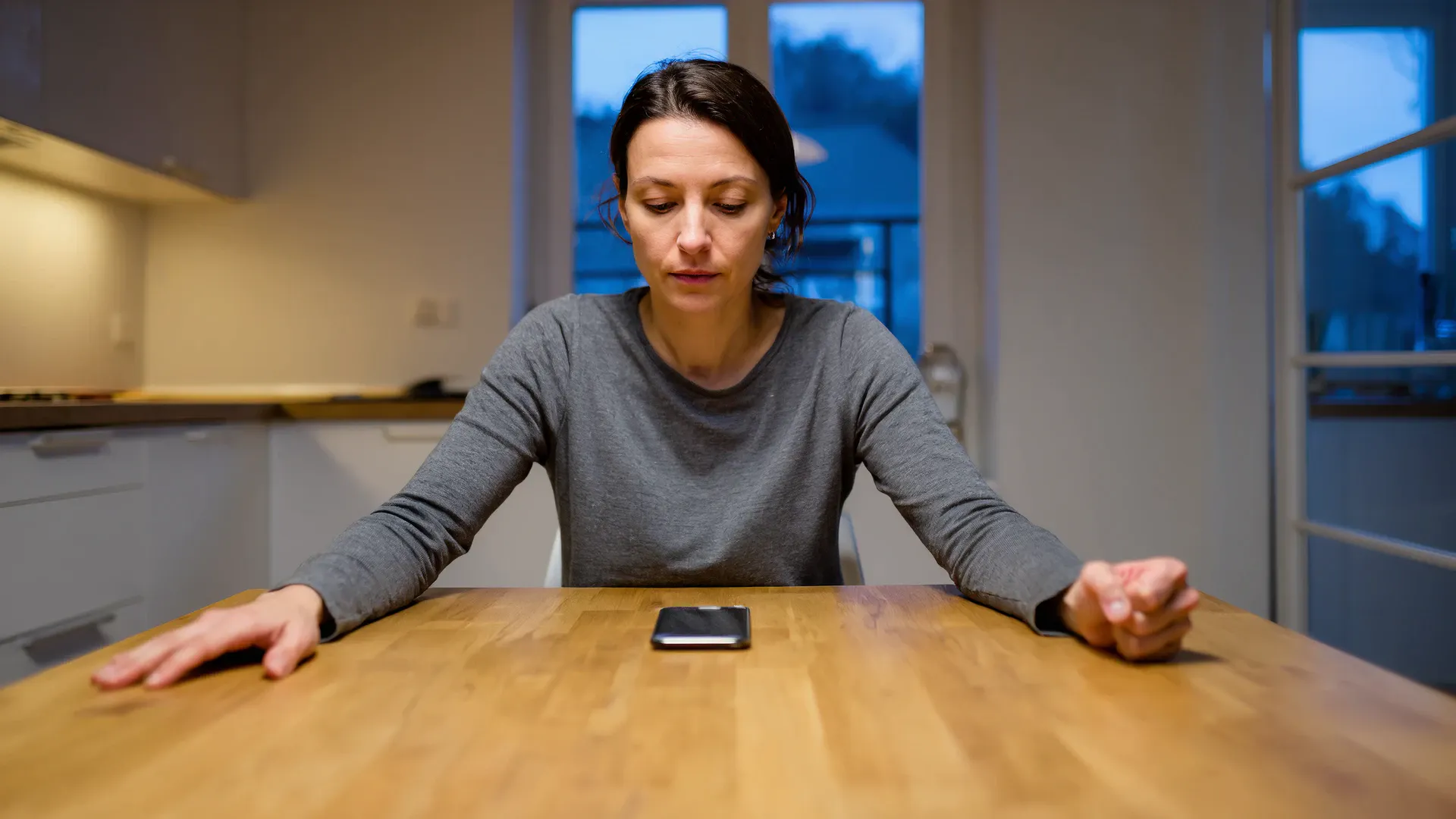 A woman sits alone at a wooden kitchen table at night, looking down with a still, blank expression. A phone lies face-down in front of her. Her left hand rests flat and loose on the table with the fingers spread; her right hand is clenched into a tight fist.