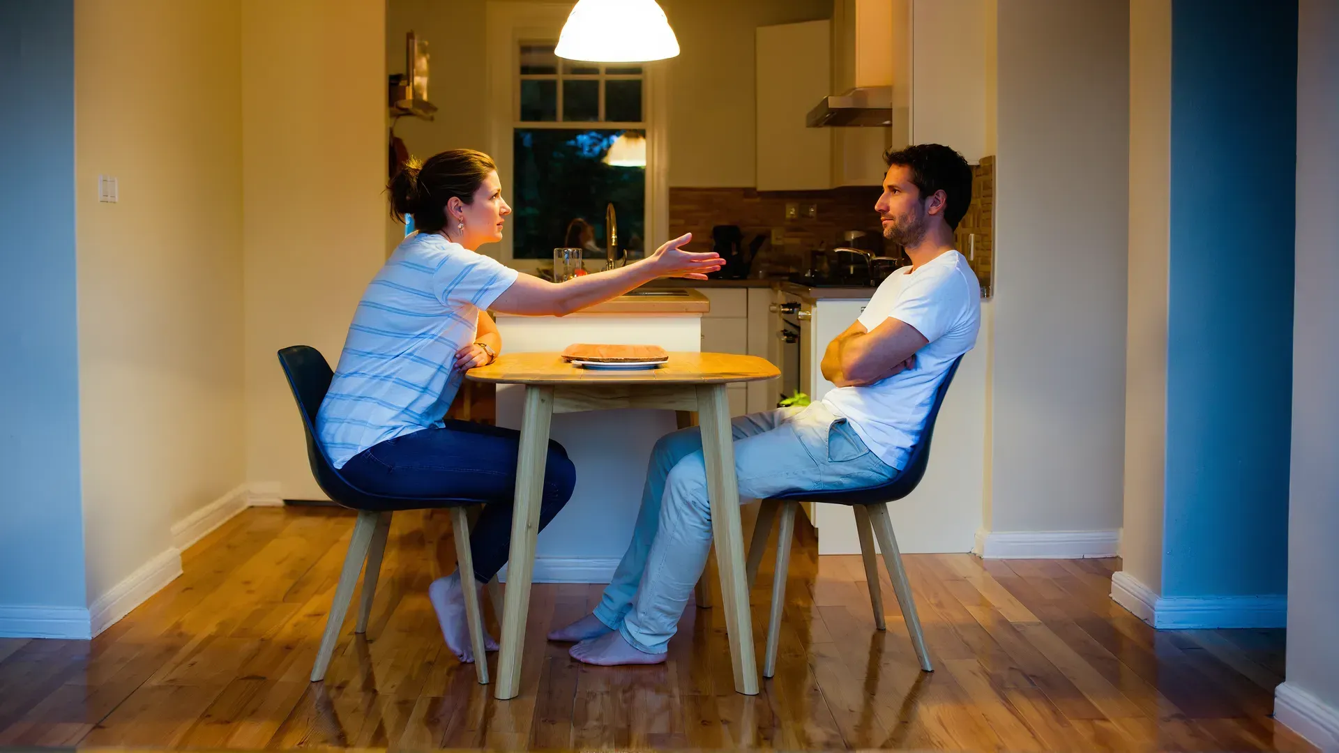 A woman reaches across a kitchen table with an open hand while a man sits back with his arms folded, turned away — one partner reaching, the other closed off.
