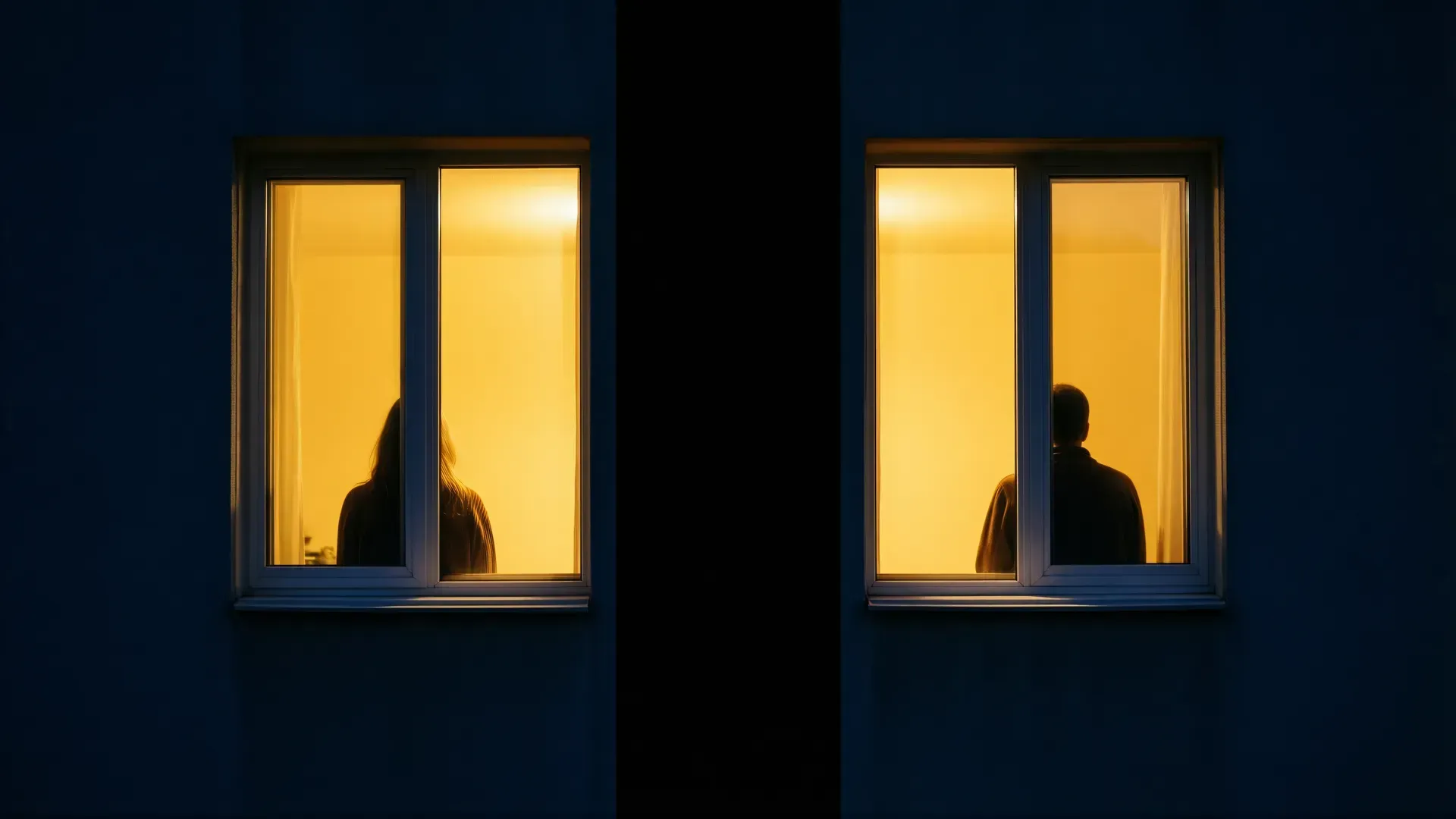 Two warmly lit apartment windows at night, a person silhouetted and turned away in each, divided by a band of darkness — two separate inner worlds that no closeness can bridge