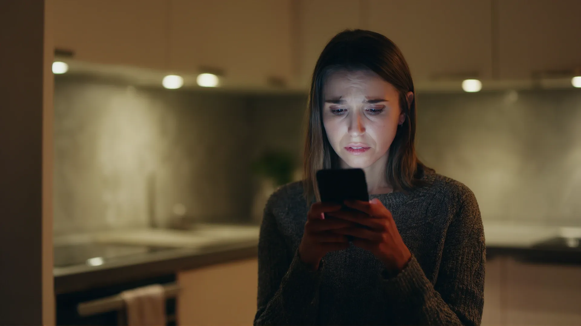 A woman stands alone in a dimly lit kitchen at night, her face lit by the glow of a phone she holds in both hands, her expression uncertain and distressed.