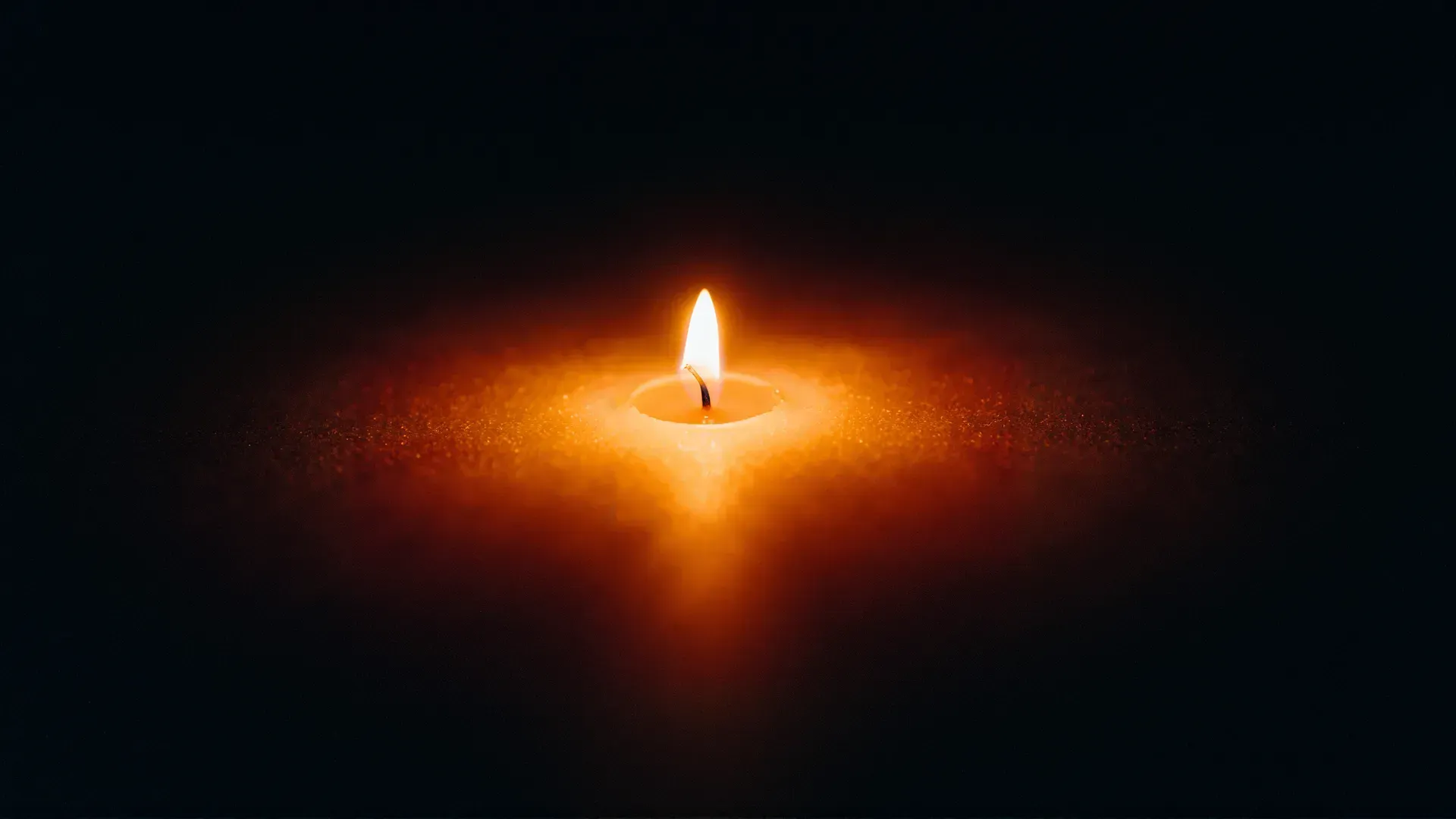 A single candle flame burning alone in deep darkness, a small contained pool of warm amber light fading to black
