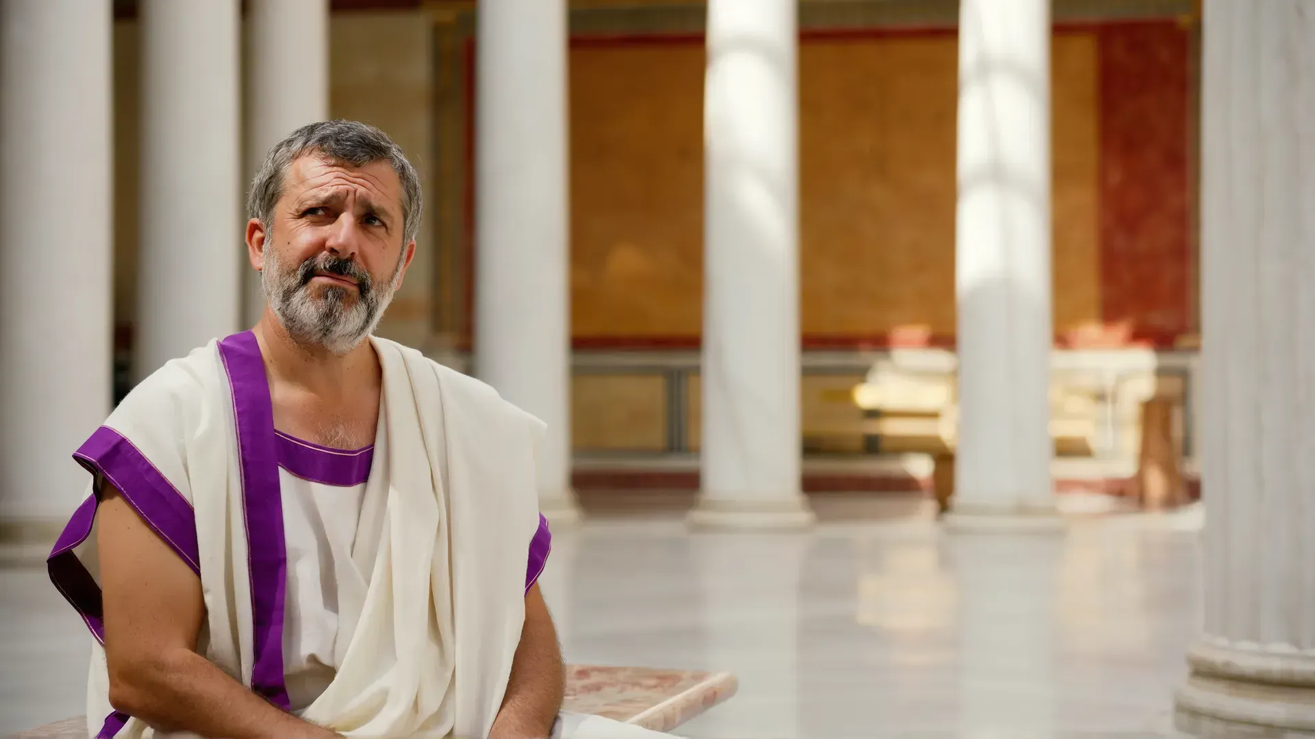 A bearded man in a white toga with a purple border sits in thought in a sunlit Roman colonnade, gazing upward as if weighing an argument.