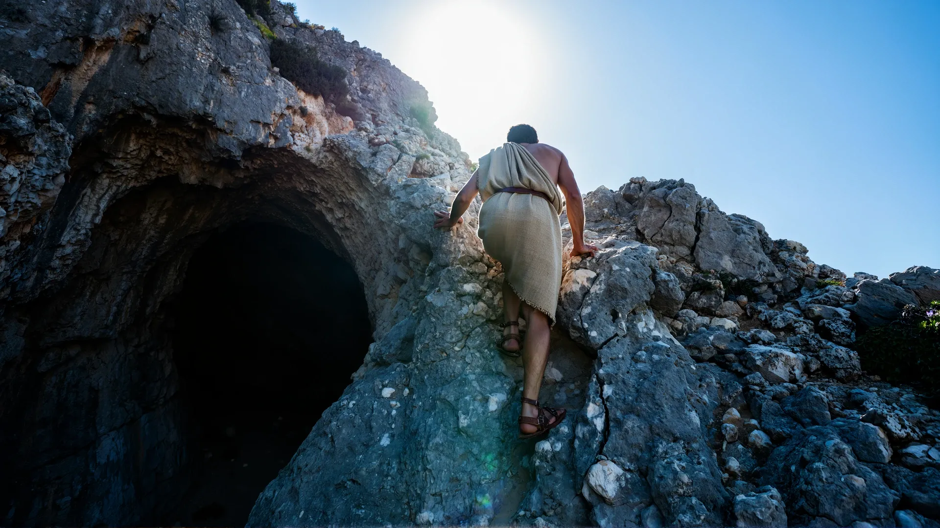 A robed figure seen from behind climbing a rocky passage out of a dark cave mouth toward a blinding burst of sunlight above — the ascent from shadow to understanding.