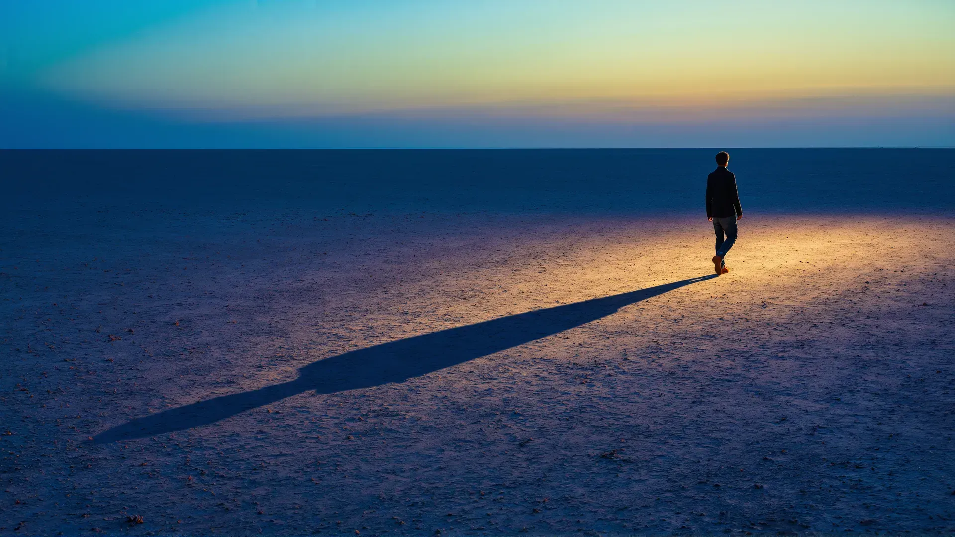 A lone figure walks across a vast empty twilight salt-flat, a long shadow trailing behind under a fading sun.