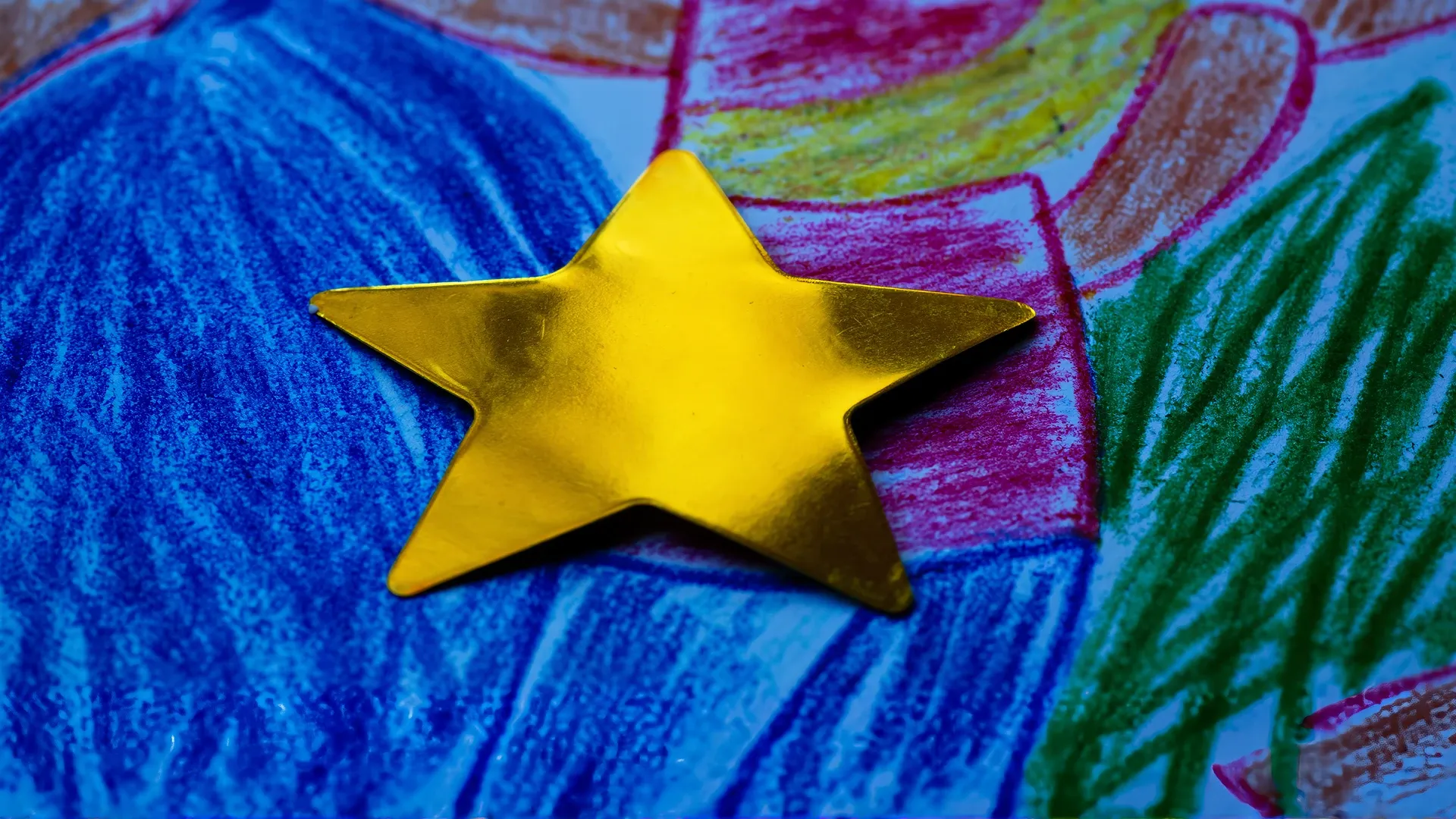 A shiny gold star sticker pressed onto a child's vivid crayon drawing, the bright star dominating and flattening the hand-drawn colour beneath it.
